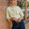 A woman leans against a red brick wall wearing a yellow quarter-sleeve shirt with a faded '21' graphic on the front, paired with dark blue jeans.