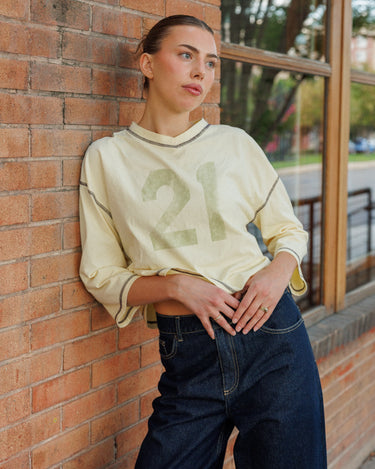 A woman leans against a red brick wall wearing a yellow quarter-sleeve shirt with a faded '21' graphic on the front, paired with dark blue jeans.
