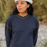 A woman wearing a navy blue quarter-sleeve V-neck sweater with gold-colored contrast stitching, paired with a white striped knit beanie, photographed outdoors against a blurred natural background.