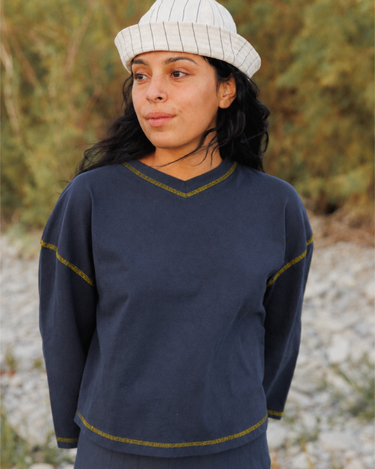 A woman wearing a navy blue quarter-sleeve V-neck sweater with gold-colored contrast stitching, paired with a white striped knit beanie, photographed outdoors against a blurred natural background.
