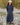 A woman stands outdoors on a rocky path with dry brush in the background, wearing a navy blue quarter-sleeve top with thin yellow piping and pleated maxi skirt, a white knit beanie, and brown shoes.