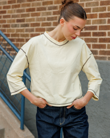 A woman wearing a yellow quarter-sleeve top with navy blue piping details on the neckline and cuffs, paired with dark blue jeans, standing in front of a brick wall.