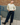 A model wearing a yellow-colored quarter-sleeve top with thin dark piping, dark wide-leg jeans, and black ankle boots, standing outdoors against a brick and concrete wall with a blue metal railing.