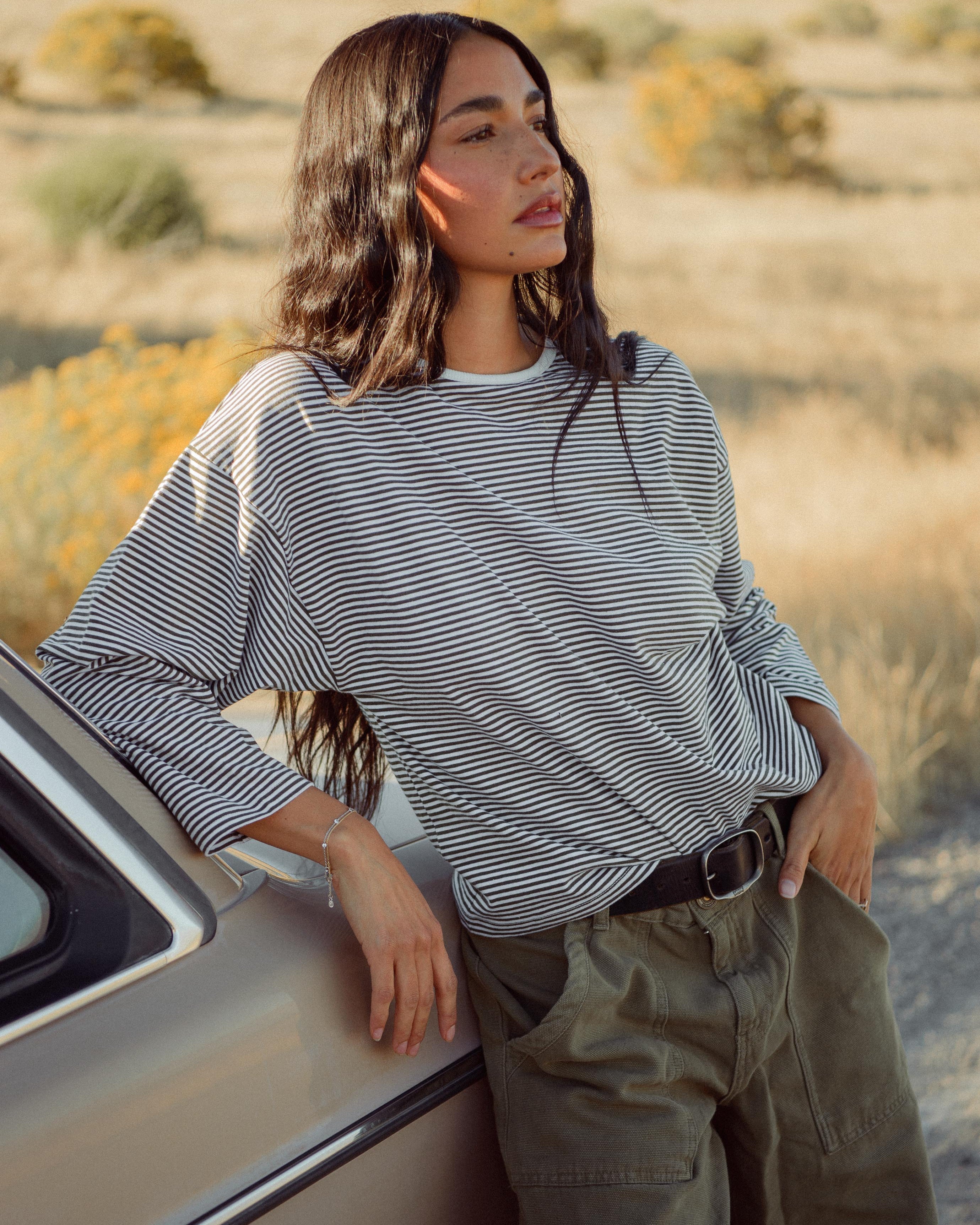 Woman wearing a striped shirt and green cargo pants, leaning on a vintage car outdoors.