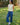 Woman wearing a light yellow sleeveless top and blue casual pants standing in a grassy area with trees in the background.