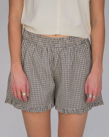 Woman wearing a pair of black and cream plaid boxer-style shorts with an elastic waistband and ruffle detail at the bottom, paired with a cream shirt.