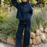 A woman stands outdoors in a garden setting wearing a dark blue cropped denim jacket with a high collar and button front, matching wide-leg denim jeans, and brown leather loafers.