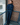 A woman wearing a dark blue denim matching jacket and jeans set stands with arms crossed, leaning against a brick wall on an outdoor metal staircase with a blue railing.