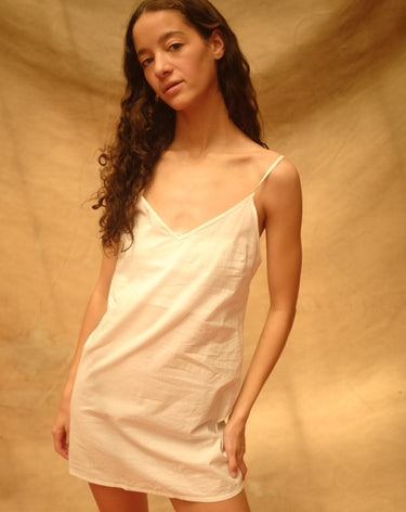 Woman wearing a short white slip dress with thin spaghetti straps and a v-neckline against a tan background.