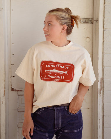 Woman wearing a cream t-shirt with a red sardine tin graphic that says "Sønderhaus Sardines", paired with denim pants.