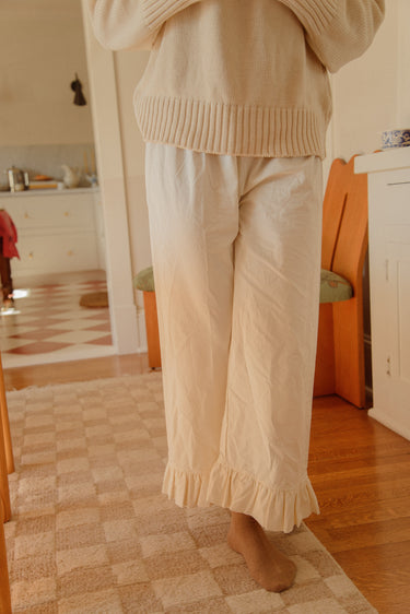 Woman standing inside a house wearing cream casual pants with a ruffled ankle hem and a cream knit sweater.