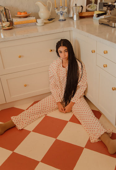 Woman sitting on orange and cream diamond-shaped tile in a kitchen, wearing a cream blouse and pants set with a red floral print.