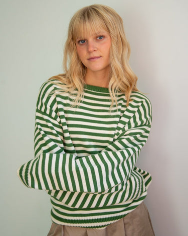 Woman wearing a green and white striped long-sleeved crewneck sweater and white pants, standing against a plain backdrop.