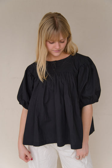 Woman wearing a black babydoll-style blouse with short balloon sleeves and white pants standing against a plain background.