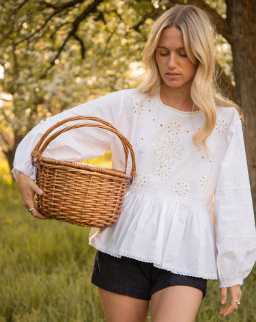 Woman holding a wooden basket in a grassy field outdoors wearing a white eyelet long sleeve blouse and black shorts.