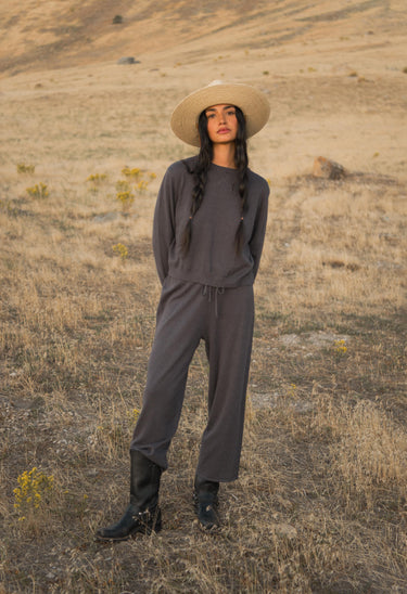 Woman wearing a wide-brimmed hat and dark outfit standing in a dry, open landscape.