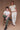 Man and woman against a brown background. The man is sitting on a wooden chair wearing a white shirt with green jeans. The woman is standing and leaning on the chair, wearing a cream collared cardigan and white jeans.