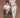 Man and woman against a brown background. The man is sitting on a wooden chair wearing a white shirt with green jeans. The woman is standing and leaning on the chair, wearing a cream collared cardigan and white jeans.