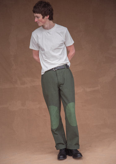 A man wearing a white t-shirt and green pants against a brown background.