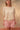 Woman wearing a beige knit short sleeve cardigan and pink plaid shorts on a tan fabric backdrop.