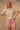Woman wearing a beige knit short sleeve cardigan and pink plaid shorts on a tan fabric backdrop.