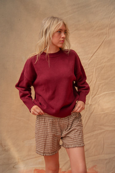 Woman wearing a merlot red knit sweater and brown gingham shorts, standing against a tan background.