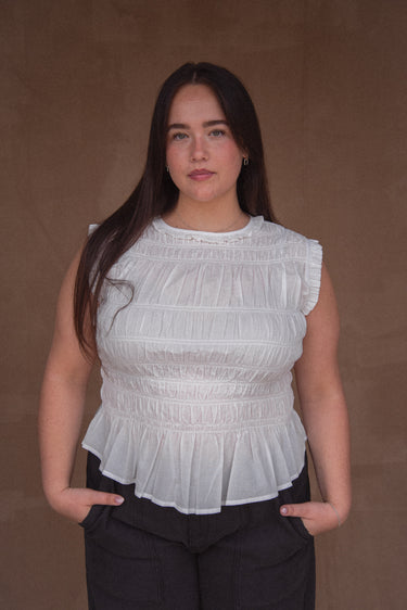 A woman wearing a white sleeveless top with a smocked bodice and ruffled hem, paired with black pants.