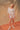 A woman wearing a white, bloomer-style shorts and a matching flowy, feminine top on a tan fabric backdrop.