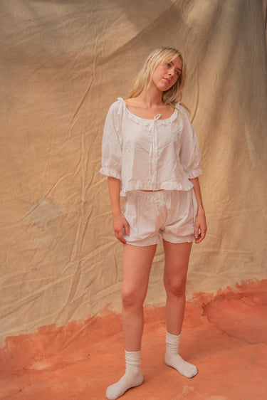 A woman wearing a white, bloomer-style shorts and a matching flowy, feminine top on a tan fabric backdrop.