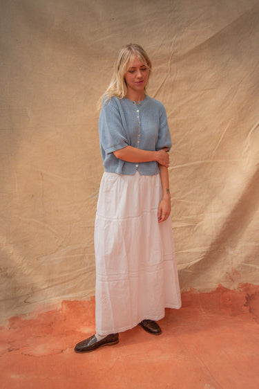 Woman wearing a blue knit short sleeve cardigan and white skirt on a tan fabric backdrop.
