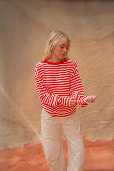Woman wearing a red and white striped long-sleeved crewneck sweater and white pants, standing against a tan backdrop.
