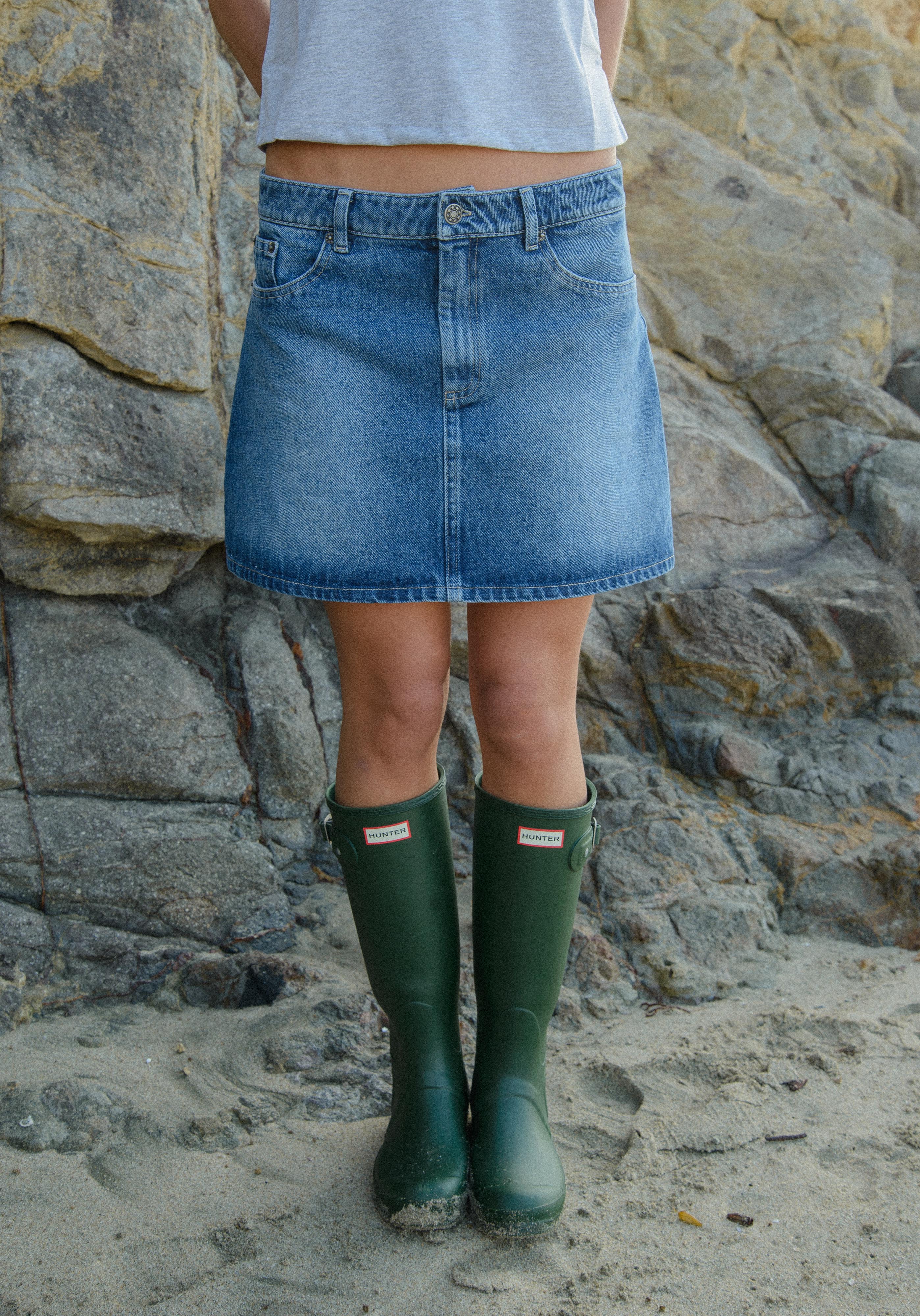 Person wearing a denim skirt and green wellington boots standing against a rocky background