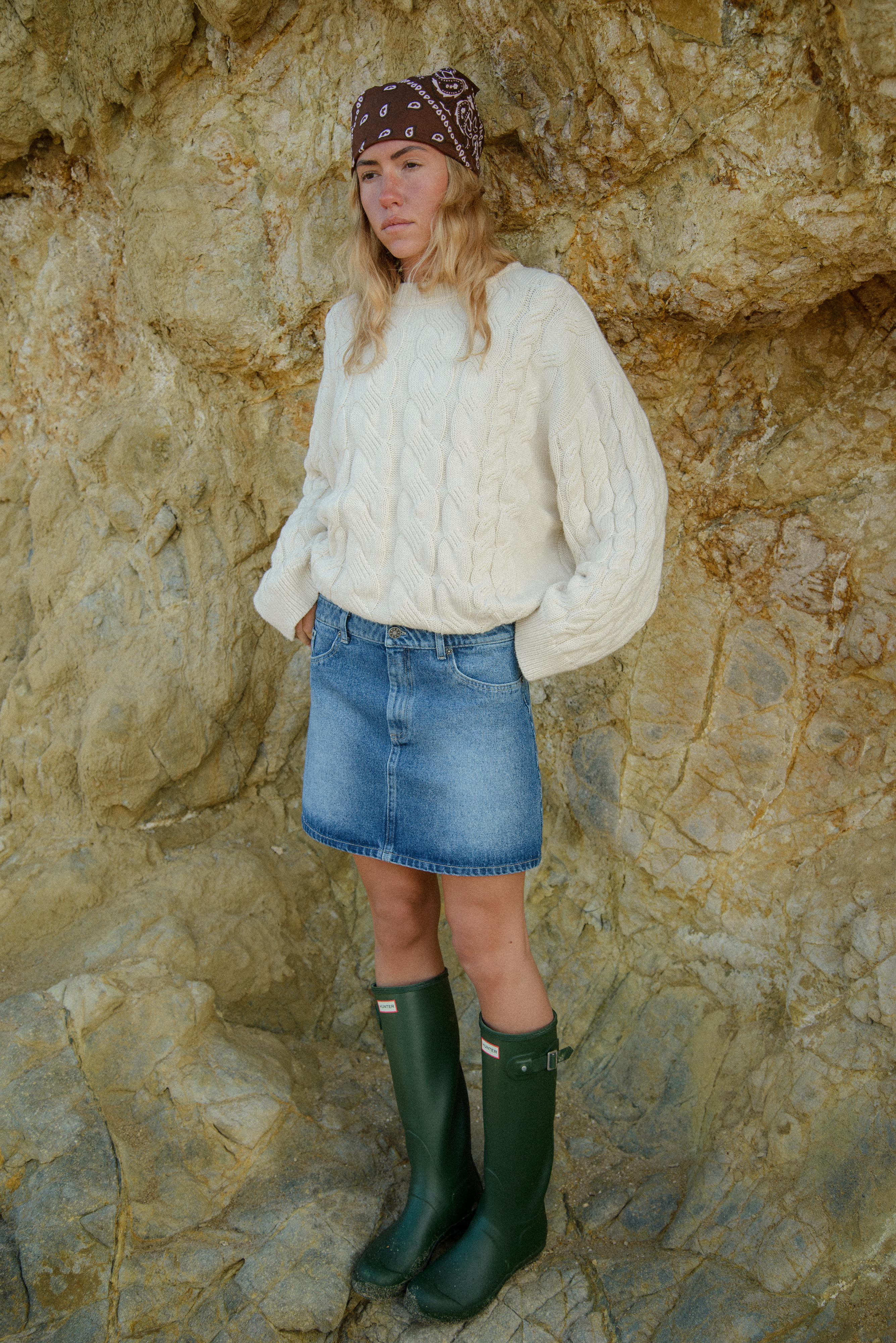 Person wearing a cream sweater, denim skirt, and green boots against a rocky background