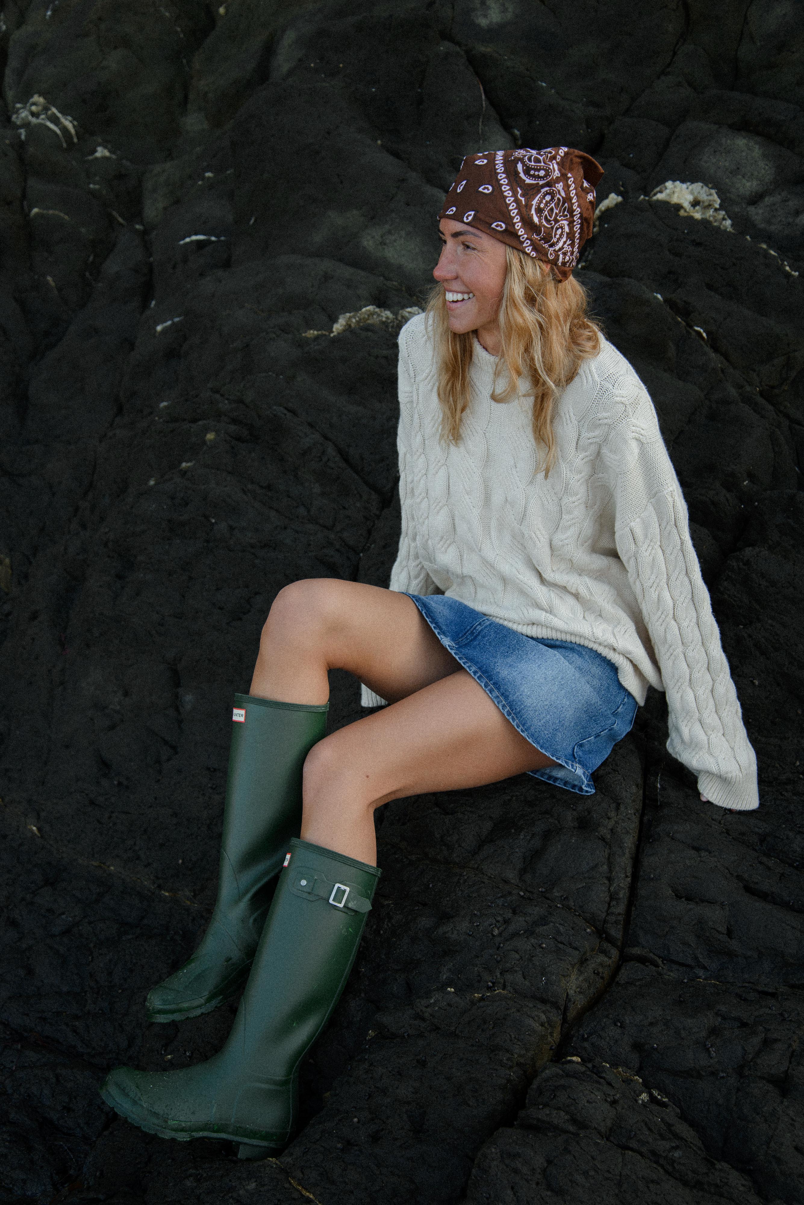 Woman sitting on a rocky surface wearing a cream sweater, denim skirt, and green wellington boots.