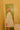 Woman standing in a bathroom wearing a white, knee-length dress with long sleeves and a ruffled collar.