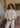 A woman standing in a room in front of a plaid couch, wearing a white long-sleeved blouse and matching pants.