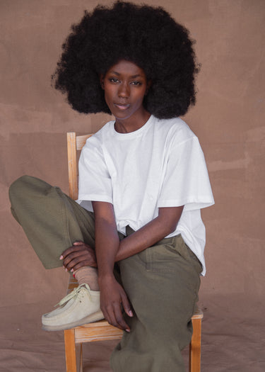 Woman sitting on a chair wearing a white t-shirt and green pants against a brown background.