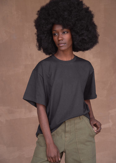 Woman wearing a navy blue t-shirt and green pants against a brown background.