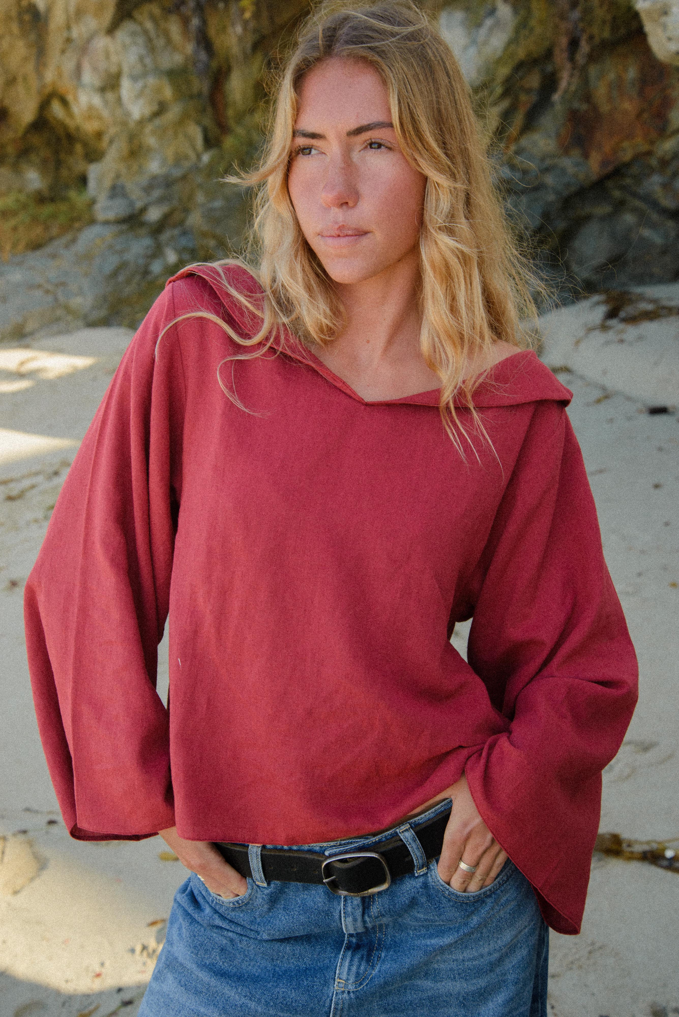 Woman wearing a red hoodie and blue jeans standing on a beach with rocks in the background