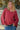Woman wearing a red shirt and blue jeans standing on a beach with rocks in the background
