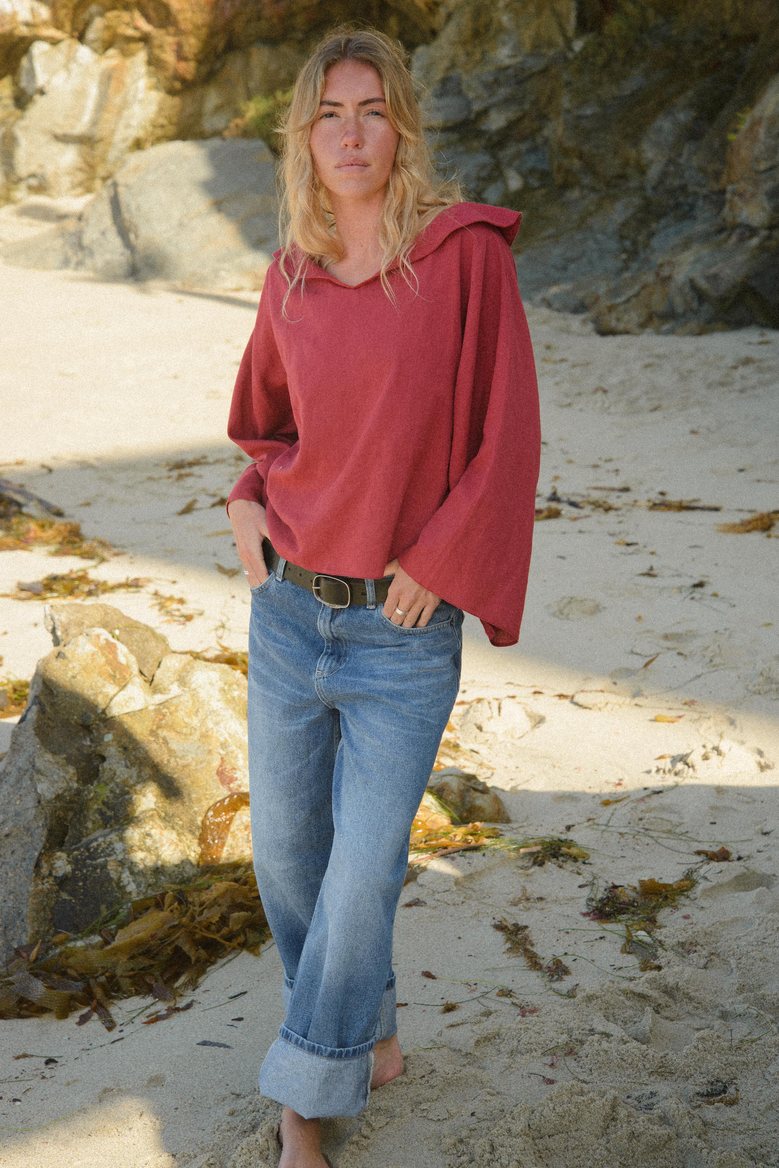 Woman in a red sweater and blue jeans standing on a beach with rocks and sand in the background.