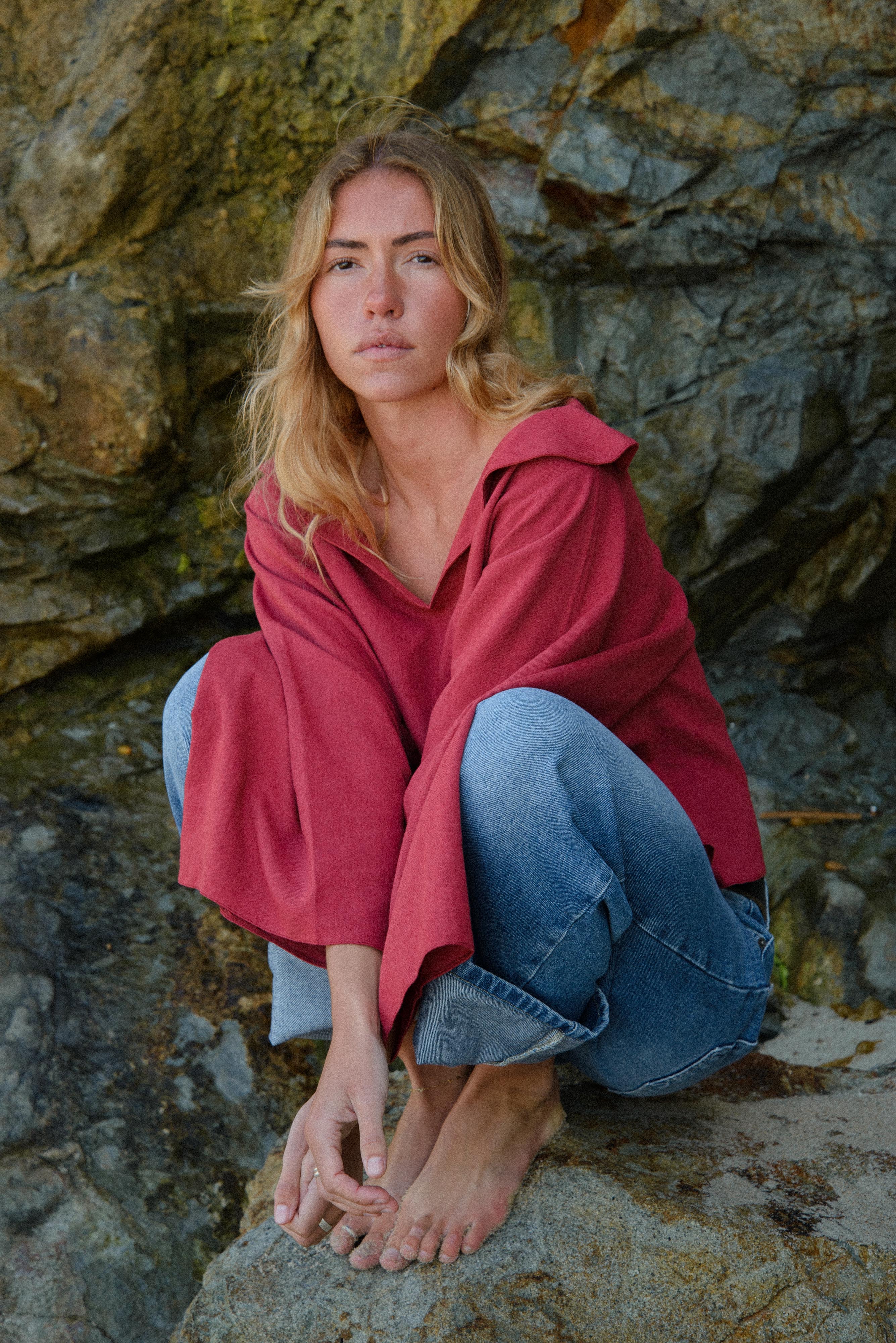 Woman in a red shirt and blue jeans squatting on rocks with a rocky background