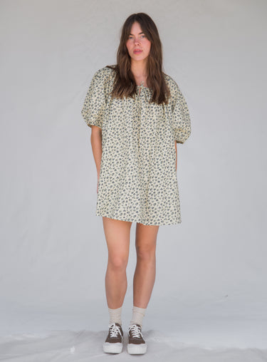 Woman standing against a white background wearing a cream floral printed, knee-length dress with a tie neckline and short sleeves.