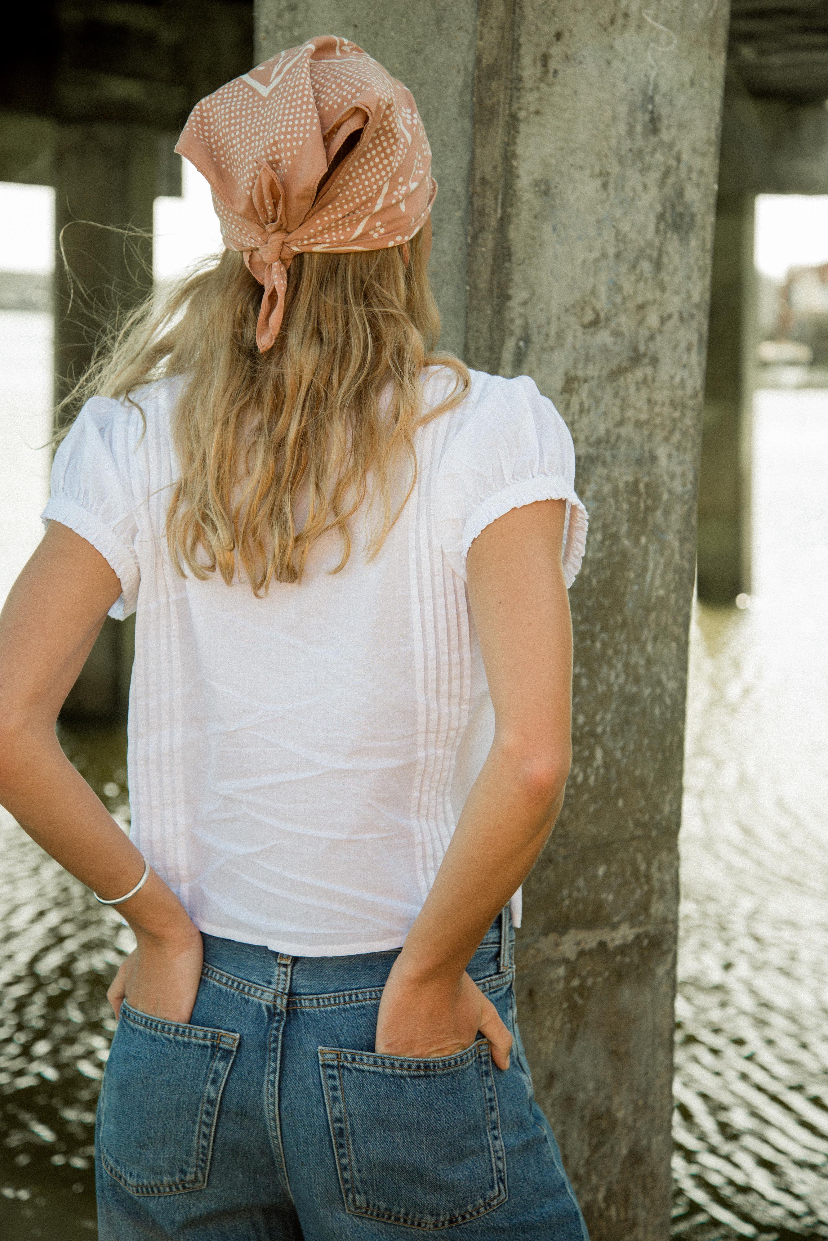 Person wearing a white top and blue jeans with a headscarf, standing by a stone wall.