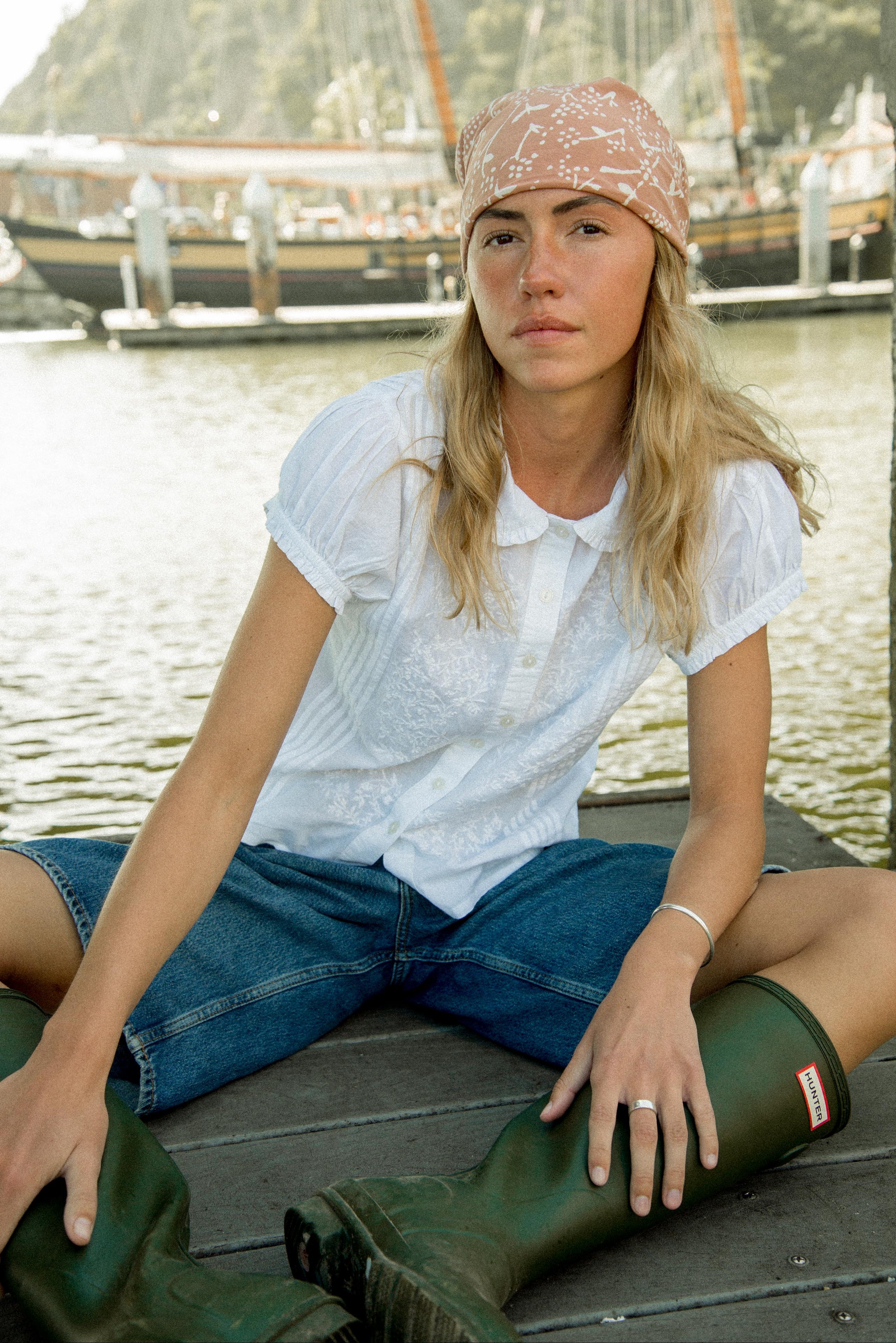 Woman sitting by a body of water wearing a white shirt, blue jeans, and green boots.