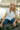 Woman sitting by a body of water wearing a white shirt, blue jeans, and green boots.