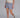 Woman wearing a pair of blue plaid boxer-style shorts with an elastic waistband and ruffle detail at the bottom, paired with a cream shirt and brown sneakers.