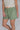 Woman wearing a pair of green plaid boxer-style shorts with an elastic waistband and ruffle detail at the bottom, paired with a cream shirt.