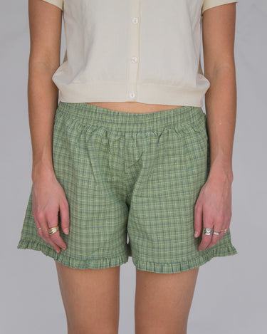 Woman wearing a pair of green plaid boxer-style shorts with an elastic waistband and ruffle detail at the bottom, paired with a cream shirt.