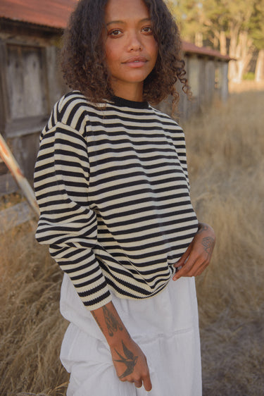 Woman standing in a field, wearing a black and white striped long-sleeved crewneck sweater with a white skirt.